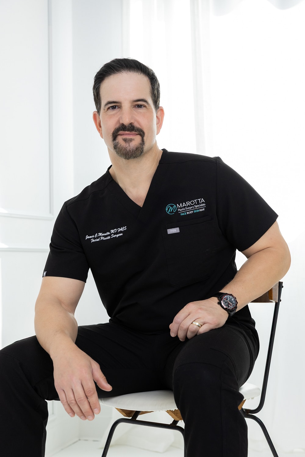 Male surgeon in black scrubs, seated confidently.